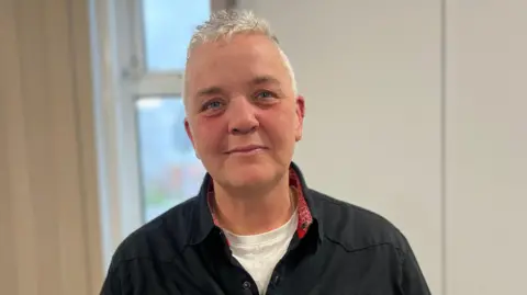 Kaz is wearing a black button shirt, the inside of the collar is red and white, the collar is open and a white t-shirt is visible underneath. They have short white hair. 