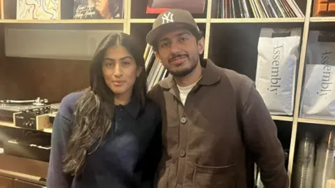 Saroop Sangha (L) and Jasbir Lalarea (R) stood in their coffee shop behind the counter. Saroop is wearing a navy jumper and has long dark hair, while Jasbir is wearing a brown cap, brown jacket and white top. 