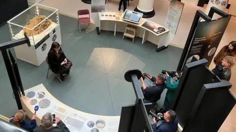 BBC/Seb Cheer A woman, Emily, sits on a chair in front of a large brown-golden mass of metal objects in a glass case. Other exhibits surround her, with photographers pointing cameras at her.