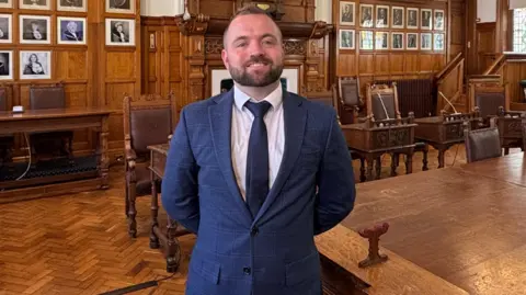 Councillor Devon Watson, who has short brown hair and a beard and is wearing a navy suit. He is smiling proudly as he stands with his hands behind his back in the Douglas Council chamber. 