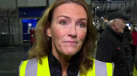 Helen O'Sullivan is wearing a yellow high viz jacket. She has wavy brown hair. Other people are standing behind her.