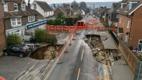 A massive sinkhole in the middle of a road, with houses to the left and right.