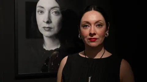 Clare Hughes at her exhibition self-portrait (in black and white behind her to the left) wearing black top, earrings and red lipstick