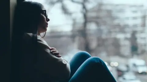 A young woman looks out of a window while sat on the sill. She has blue trousers and wears a white top with her hands folded across her chest. She has dark hair past her shoulders and wears glasses.