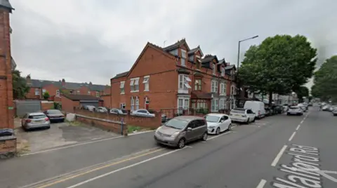 Row of red-brick terraced houses on a residential street with parked cars along the curb, a large leafy tree on the pavement, and overcast sky above.
