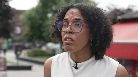 A woman with curly black shoulder length hair and colourful glasses, wearing a sleeveless cream top, is interviewed standing in a London street.