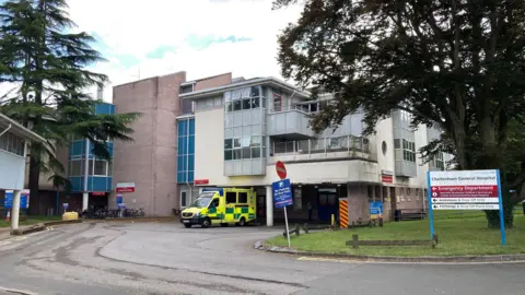 The exterior of Cheltenham General Hospital in Gloucestershire. It is a largely grey building with some cream panels, and there is one ambulance parked outside