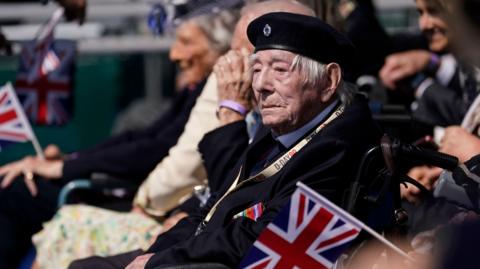A Normandy veteran sat in a wheelchair attending the UK's national commemorative event for the 80th anniversary of D-Day