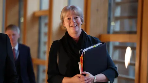 Shona Robison carrying a black folder as she heads into the Scottish Parliament to deliver the budget. She is smiling and wearing a black outfit. 