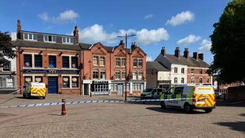Police cordon at Market Place in Ilkeston with three police vans parked on a paved area next to a row of red-brick buildings