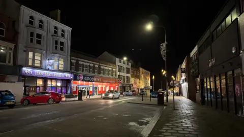 A photo of Prince Street in Bridlington at night. Street lights illuminate the street. Cars are parked on one side. Shops and bars can be seen.