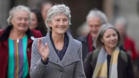 PA Media Catherine Connolly wears a woven blazer. She has short cropped grey hair and is waving and smiling.