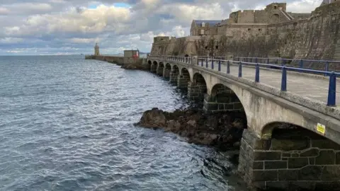 BBC A long multi-arched bridge which leads to a lighthouse and a castle with the sea to its left. The sea is blue. The sky is blue with white clouds. 