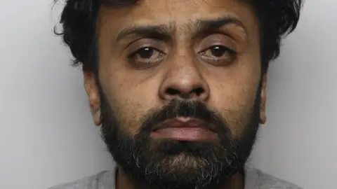 A polie mugshot of Romon Haque, who has thick unkempt black hair and a black beard and is wearing a grey top, and stares blankly into the camera.