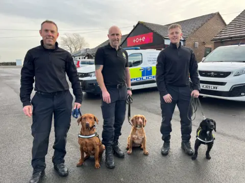 Three male police dog handlers standing with three dogs on leads in front of a police van. The men are wearing black uniforms. The dogs are sat on the ground. From left to right: a red labrador, red cocker spaniel and black cocker spaniel.