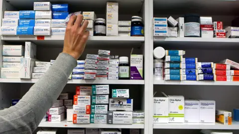 PA A person in a gray jumper reaches for medicine on a pharmacy shelf. The shelves are stacked with boxes and bottles of various medicines.