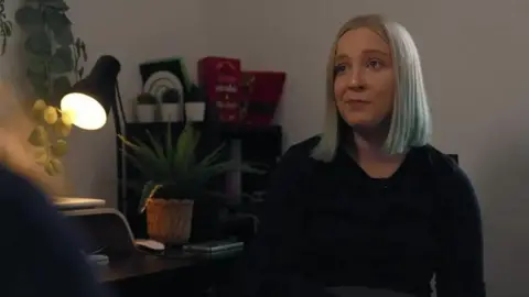 Stefanie Harman sat by her desk in her home talking to the BBC. She grey-white hair in a bob and is wearing a black top.