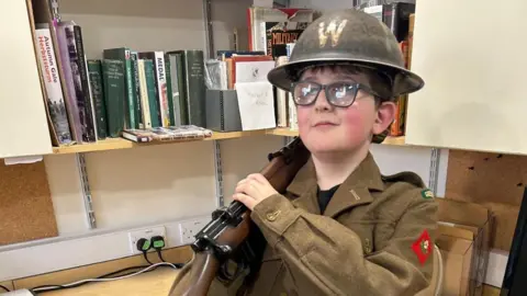 Family handout Ben, wearing a World War Two soldier's uniform, holding a gun. He is in a room with books on shelves. He has on glasses. 