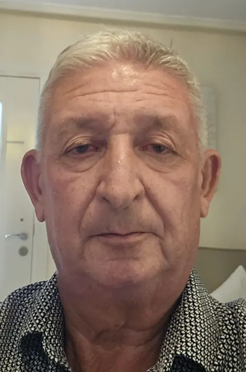 Mike Heighway A man with short white hair looks into the camera without smiling. He is clean shaven and wears a white shirt with a dense black pattern on it.