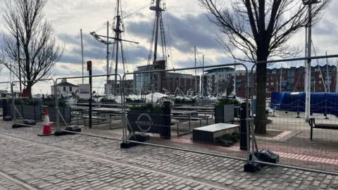 A cobbled street with a tall metal barrier fencing off trees and a pedestrianised walkway. In the distance are boat masks and behind them is a row of tall flats and commercial buildings.