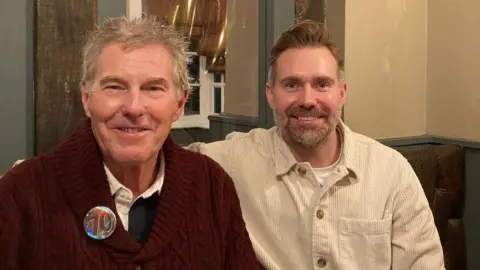 An older man and a younger man smile at the camera. The man on the left is wearing a burgundy jumper and has grey hair. The man on the right is wearing a cream-coloured corduroy shacket and has brown hair and a short beard.