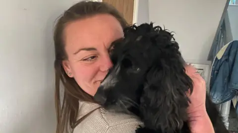 Rachel Douglas is a woman in her 40s with long brown hair and wearing a brown top. In her arms is a black spaniel