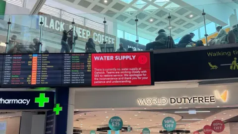 A red sign alerts passengers to the water supply issue on a screen in a terminal building. A World Duty Free sign is underneath and a pharmacy sign is nearby. A further screen shows flight information.