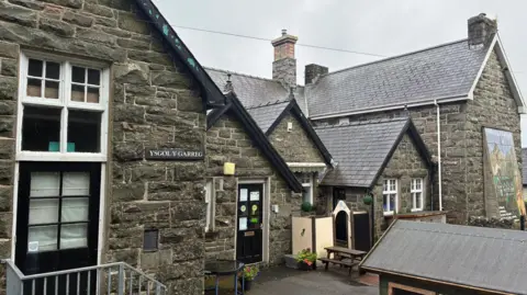 General view of Ysgol Y Garreg, a stone building with black doors and white windows - the building is in the traditional school format of gabled ends and a central section fronting onto a small yard