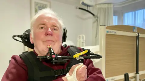 BBC A man with thinning white hair and a maroon T-shirt is pictured with a back headrest, possibly connected to a wheelchair, and has a black chest harness that clips together at the front. He is pictured in front of a raised, hospital-style bed.