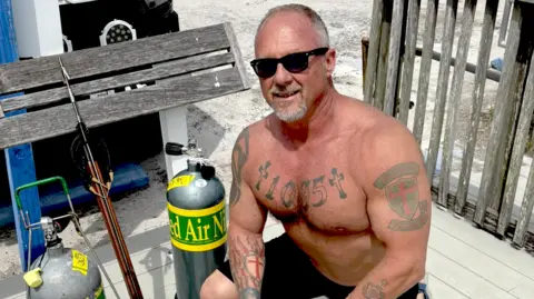 Facebook Johnny Mulford, a man with greying hair cut short and a greying goatee beard, crouching down with his shirt off so his tattoos are visible. He is wearing shorts and sunglasses and is kneeling beside some tanks of what appear to be air and what appears to be a harpoon.