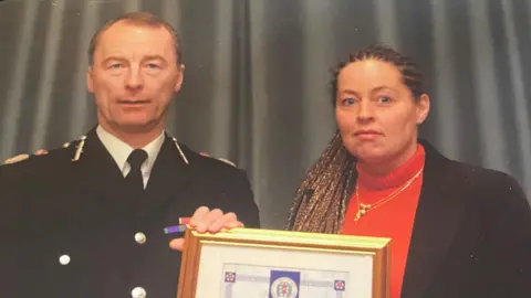 Whendie Backwell Whendie Backwell, a woman with brown hair in cornrows and plaited into long braids holds a certificate in a gold frame. She is wearing a red jumper. Next to her is an older man with thinning grey hair, possibly high up in the police force. He is wearing a suit.