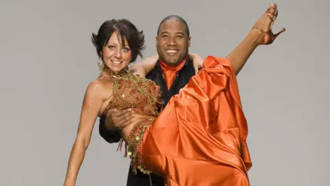 Nicole Cutler with short dark hair and wearing an orange satin and sparkly dress being held with a foot in the air by John Barnes who is wearing a black suit and orange cravat.