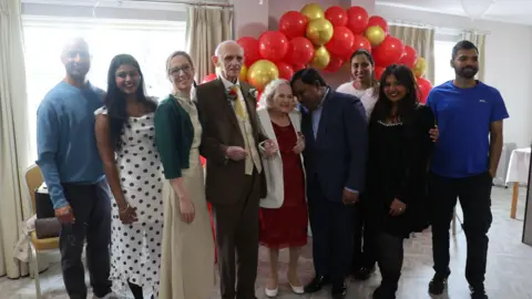 Charlton Grange Care Home Nine people (including the bride and groom and care home staff) posing for a picture in front of some red and gold balloons.