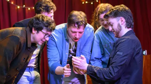 Bristol Improv Theatre Five young people crowd around a phone as they act on stage 