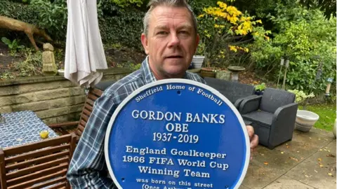 Sheffield Home of Football A grey haired man sits on a wooden chair holding up a large blue plaque to the camera