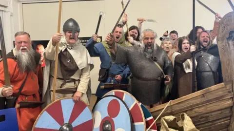 Group of people dressed in Viking costume with red and blue round shields, axes and knives. They are all looking angry with their mouths open shouting.
Their outfits are different colours, orange, tan and grey with one bearded man wearing chain mail.