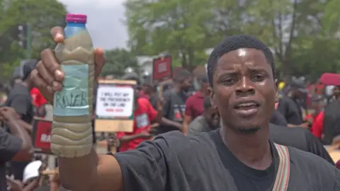 A protester wearing a black T-shirt holds up a dirty water bottle. Others can be seen behind him