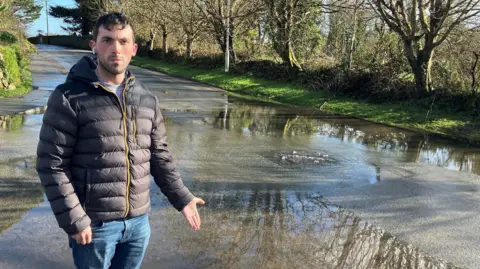 Luke Dunning is standing on a pavement and is pointing to the flooded road. There is a drain from which water is bubbling out. He is wearing a puffa jacket and has one headphone in.