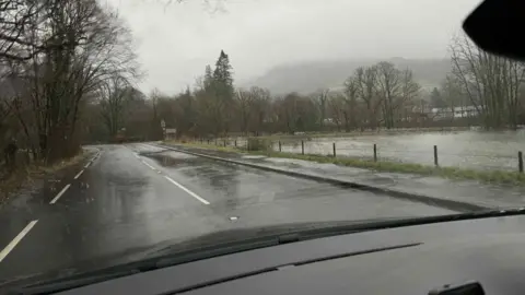 The A593 road near Rothay bridge. There is an area of green grass to the right which is covered in dark-coloured flood water. The skies are grey and gloomy and it is raining. 