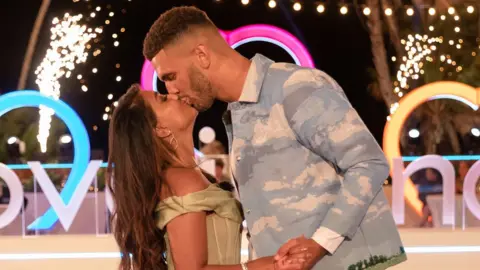 Shutterstock Sanam Harrinanan and Kai Fagan appear on 'Love Island: The Reunion' TV Show, Series 9 on19 Mar 2023. Sanam, a 25-year-old British Asian woman, kisses Kai, also 25. Sanam has long brown hair worn loose and wears a pale green off the shoulder corset-style dress. Kai, about a foot taller than Sanam, has short afro hair and wears a denim jacket, his hand on her waist. They're pictured at the Love Island villa at night, with festoon and neon lighting