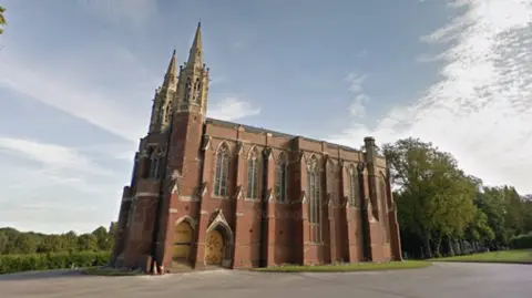 Google A brick cemetery building with arched windows and doors, and two spires at one end. It is surrounded by trees and green space.