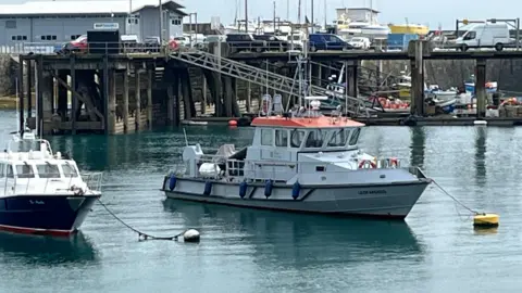 BBC Photo the Leopardess, a great boat with an orange roof and blue buoys over the side. It is docked in a harbour next to a second boat just out of frame.