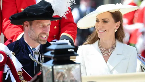 The Prince and Princess of Wales in a horse carriage. The prince wears ceremonial robes and hat, the princess is in a white jacket and white hat with a pearl necklace.
