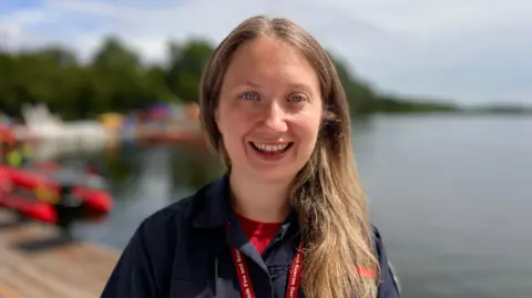 Martin Giles/BBC A head and shoulders shot of Lucy Goldsmith. She has long light brown hair pulled onto her left shoulder, over a navy shirt, which has a red T-shirt beneath and she is smiling. Behind her is a blurred-out expanse of water, trees, powerboats and sky.