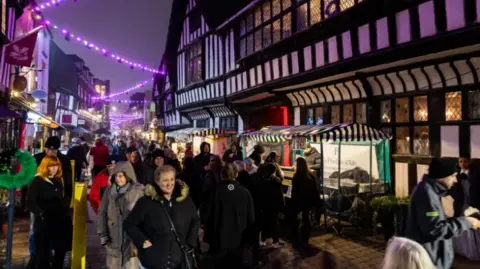 A crowd of people walk through a street with market stalls lining the path and purple lighting above them.