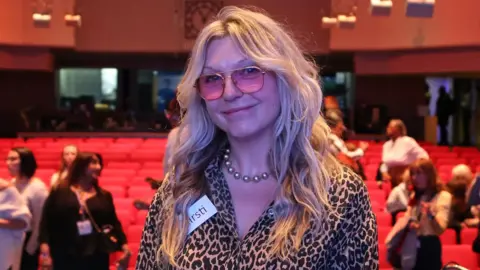 BBC / Woman's Hour Kirsti wears pink-tinted glasses and a leopard print shirt. She is smiling into the camera.