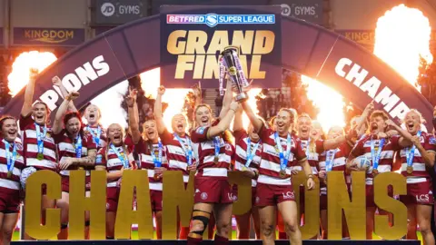 Wigan Warriors' women's team lift the 2025 Super League Trophy, with the word 'CHAMPIONS' in gold letters in front of them