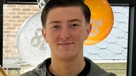 An 18-year-old man standing in front of orange, white and black balloons by a window. He has short dark hair and is smiling and is wearing a blue jacket and a black top underneath.