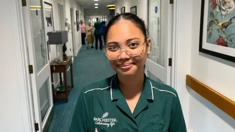 John Devine/BBC Khaye Rieta, 22, has dark hair pulled tightly over her head and is wearing glasses with a light frame. She is wearing a green overall top with white piping. Behind her is a corridor in a care home, green carpet and white walls, some residents and staff can be seen in the distance.