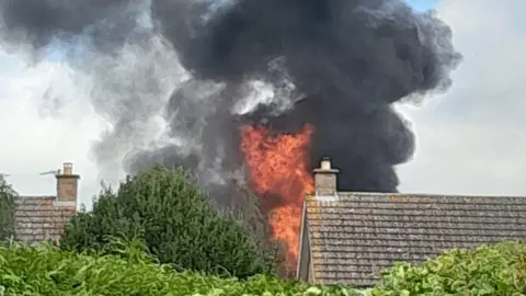 Nicky Hobson Flames and smoke rise behind rooftops. 